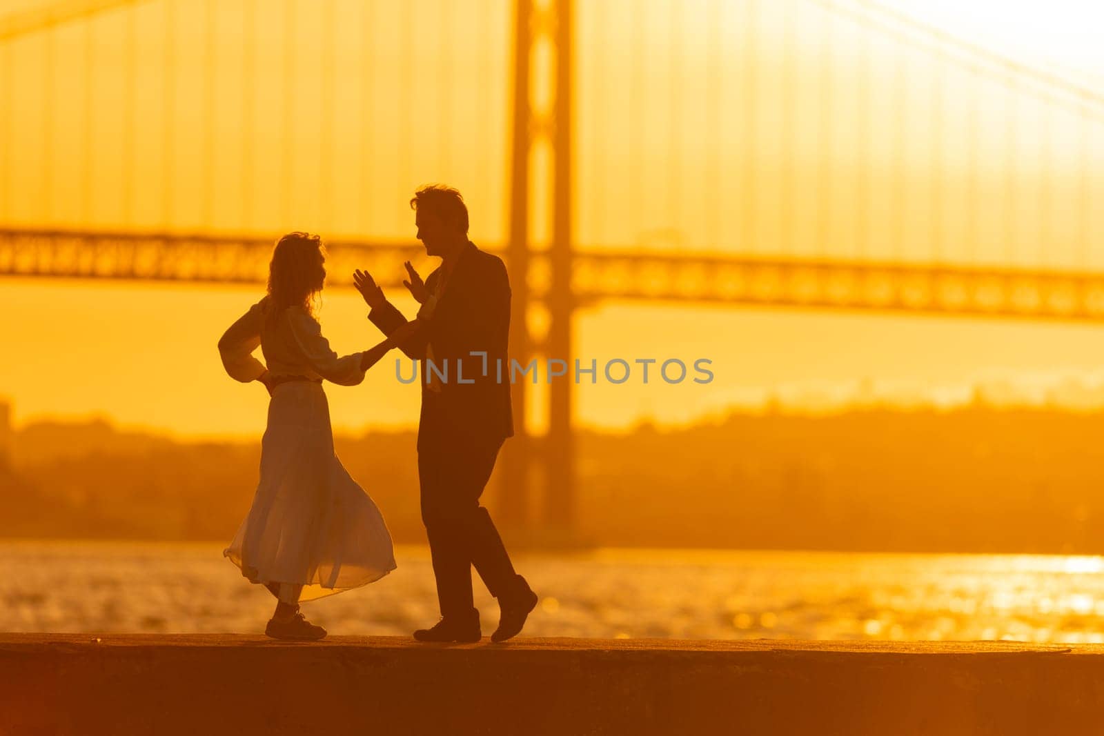 A silhouette of a couple dancing on a pier as the sun sets behind a bridge. The golden light of the sunset creates a romantic atmosphere.