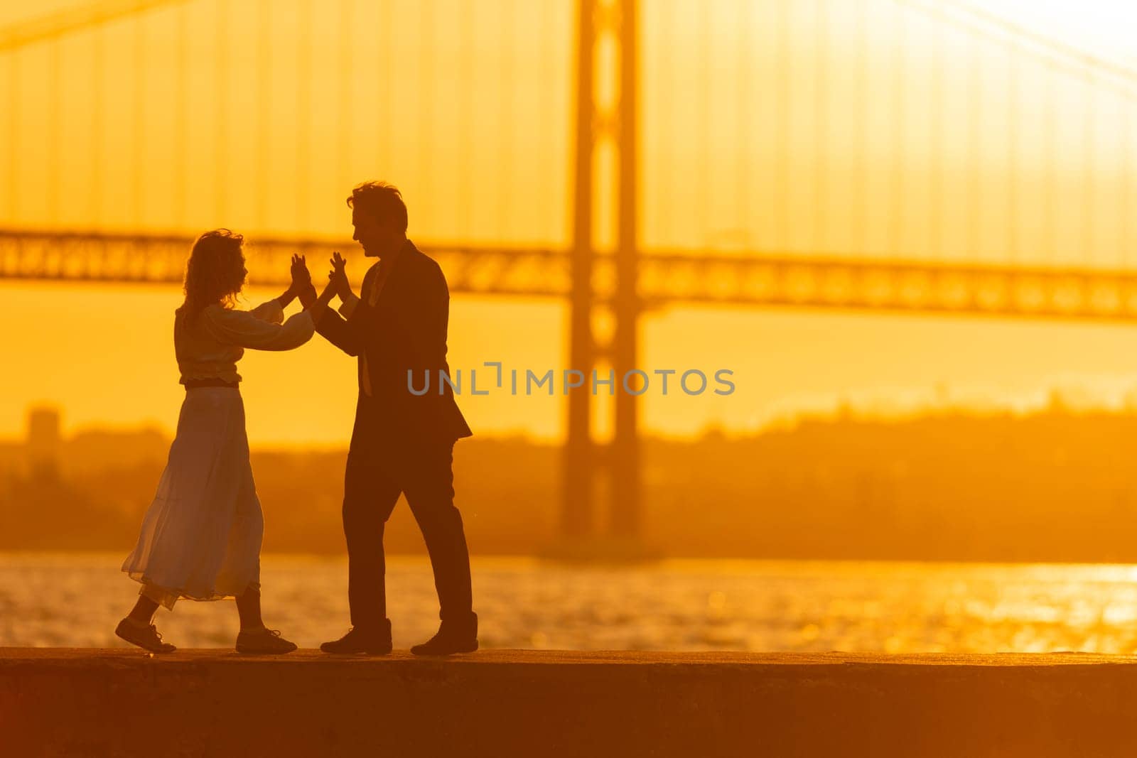 A silhouetted couple dances together on a waterfront as the sun sets behind a bridge.