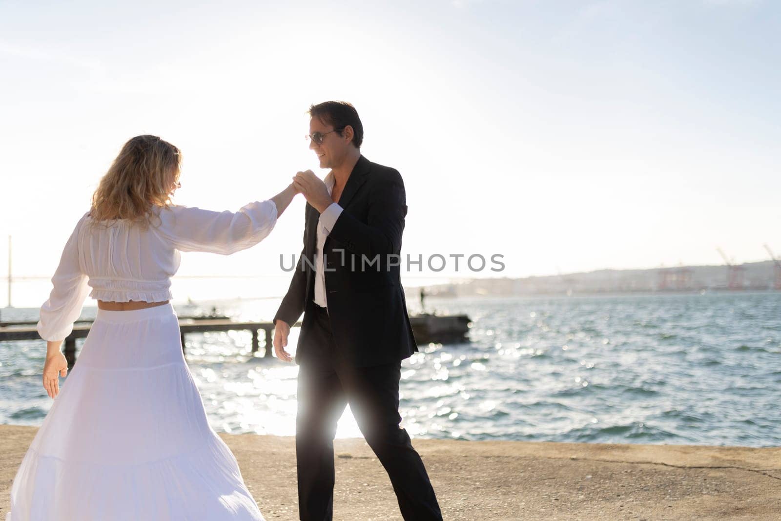A woman in a white dress and a man in a suit dance by the water as the sun sets in the background.