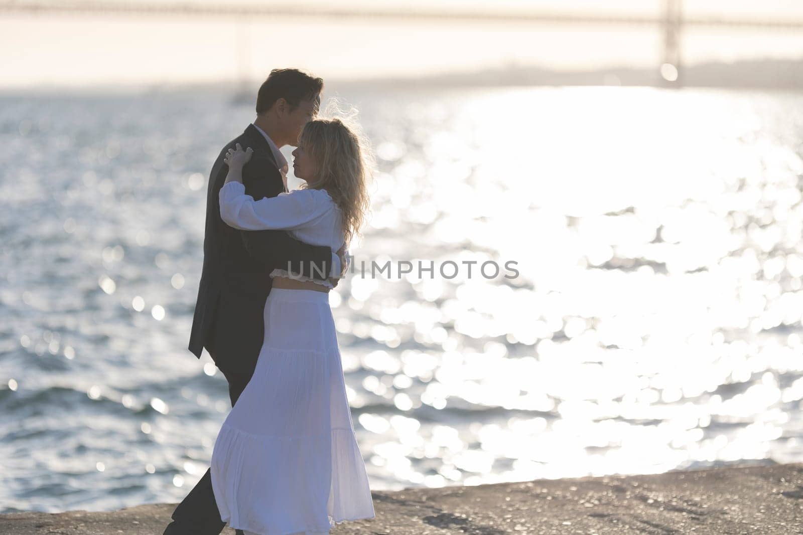 A couple dances on a rocky shore as the sun sets behind them, creating a warm glow over the water.