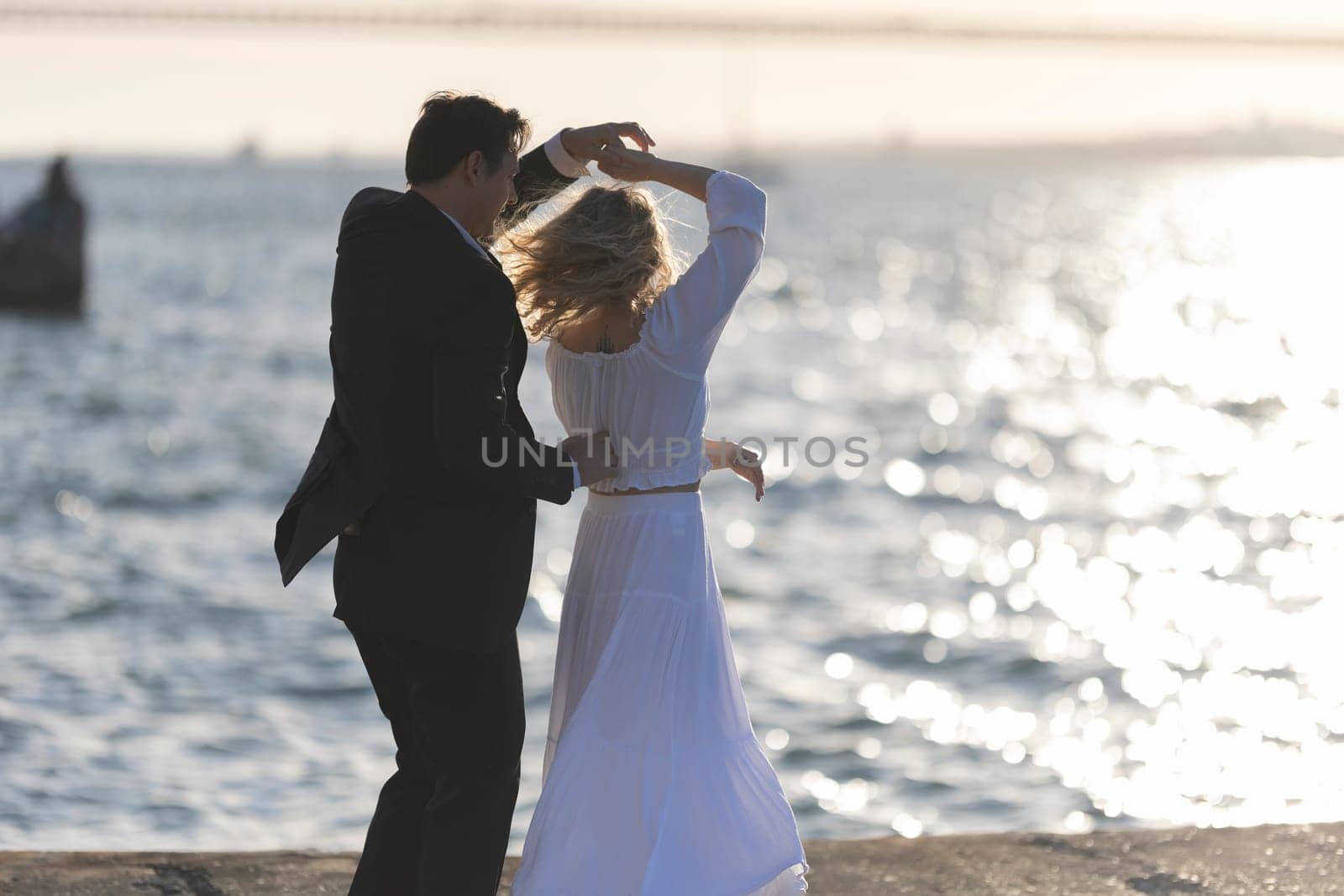 A woman in a white dress dances with a man in a suit, silhouetted against a golden sunset over the ocean.