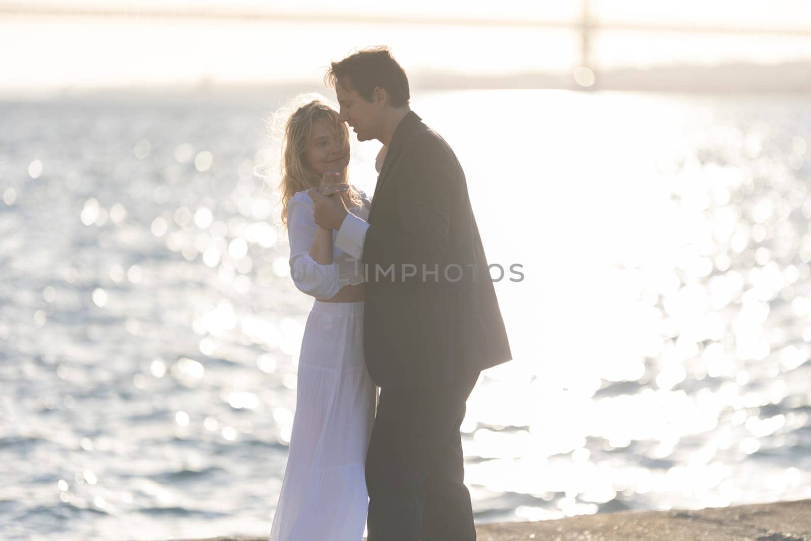 A man and woman dance together by a body of water, with the setting sun casting a warm glow.