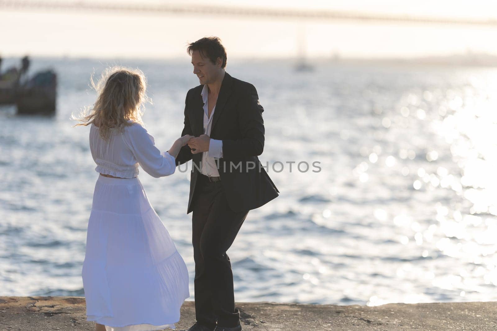 A woman in a white dress dances with a man in a suit, smiling as the sun sets behind them and casts a golden glow on the ocean.