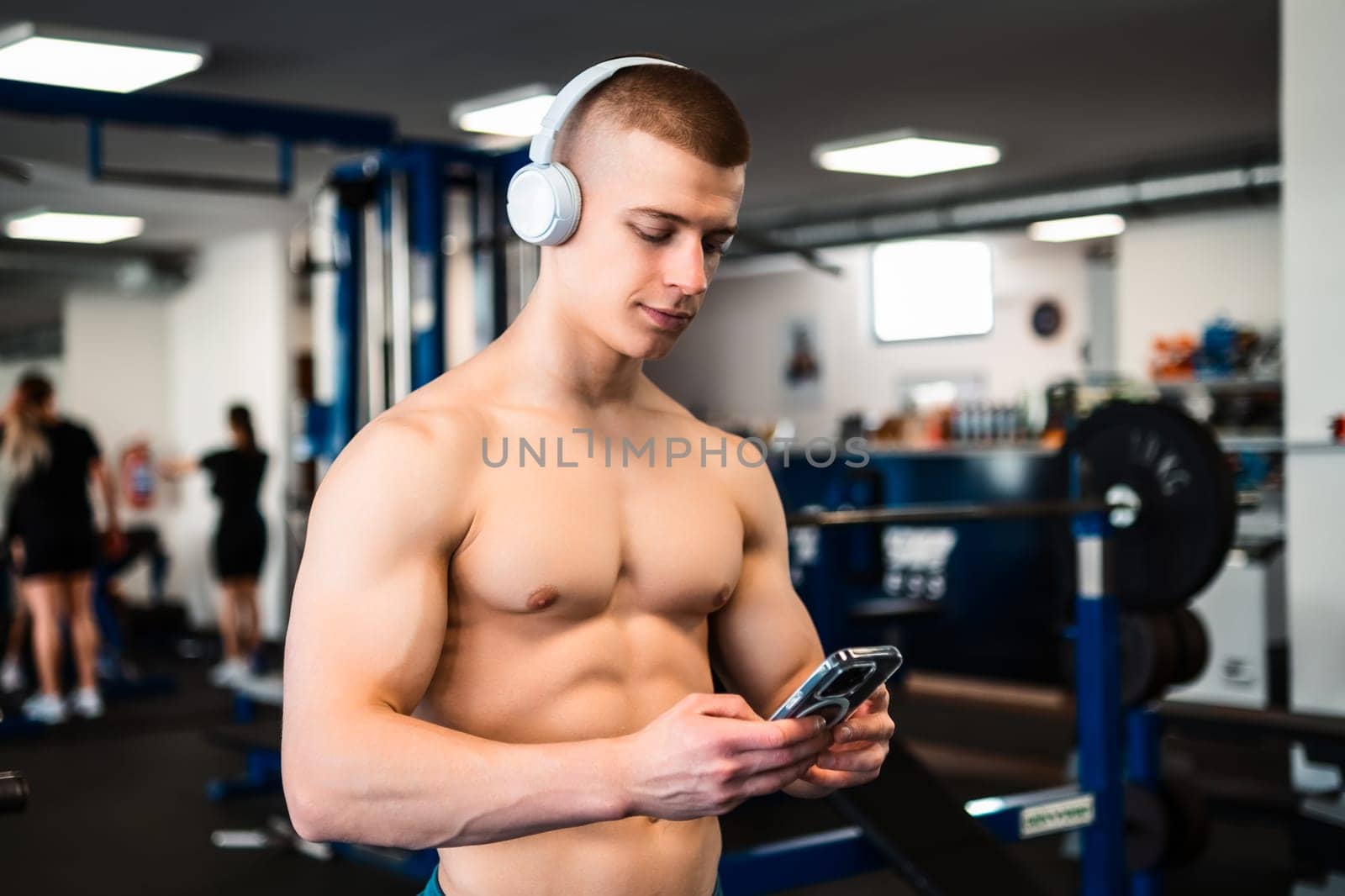 A shirtless man listens to music while wearing headphones at the gym.