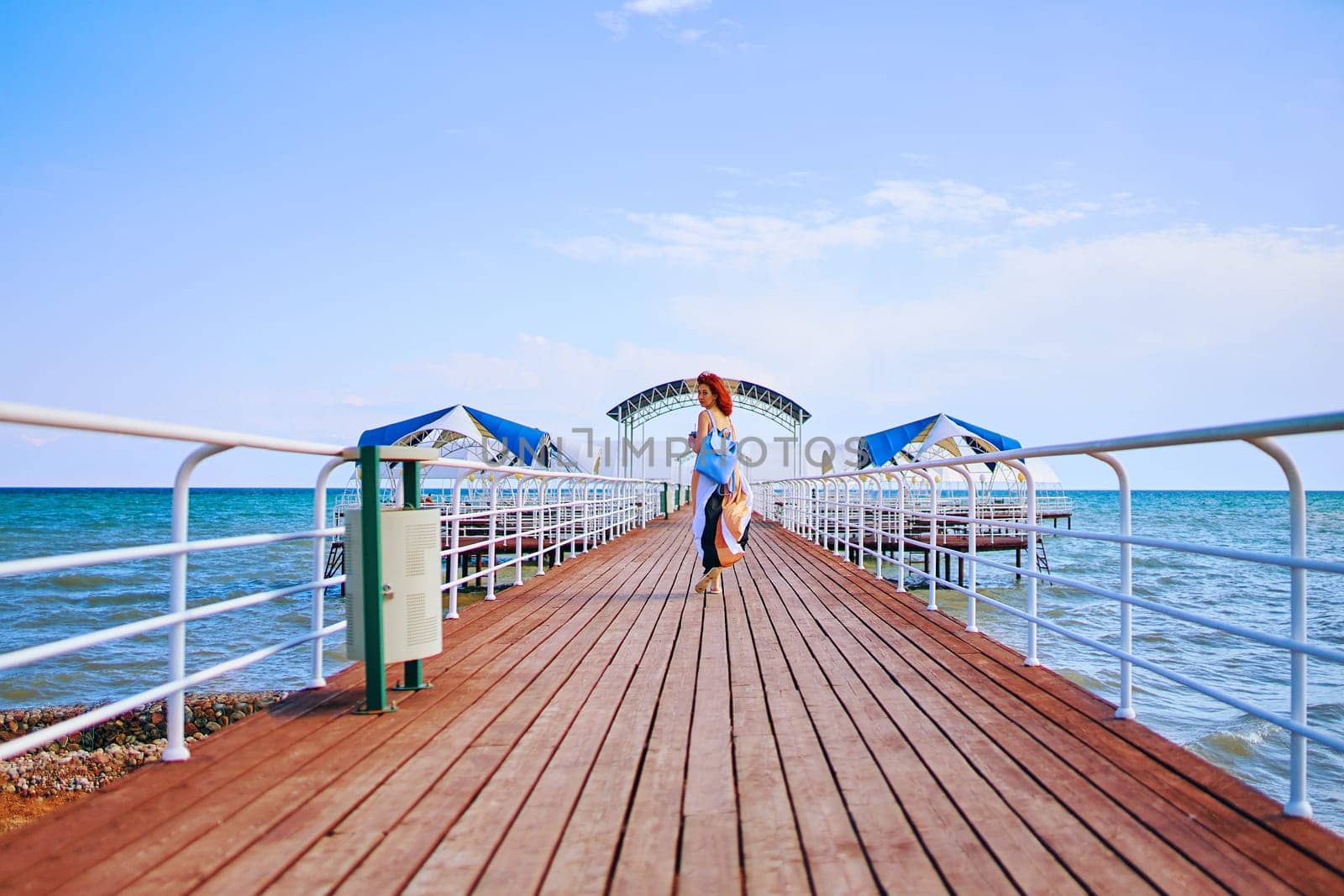 With the sea as a captivating backdrop, a red-haired woman in a summer dress and backpack graces the resort pier with her presence, creating a scene of timeless beauty.