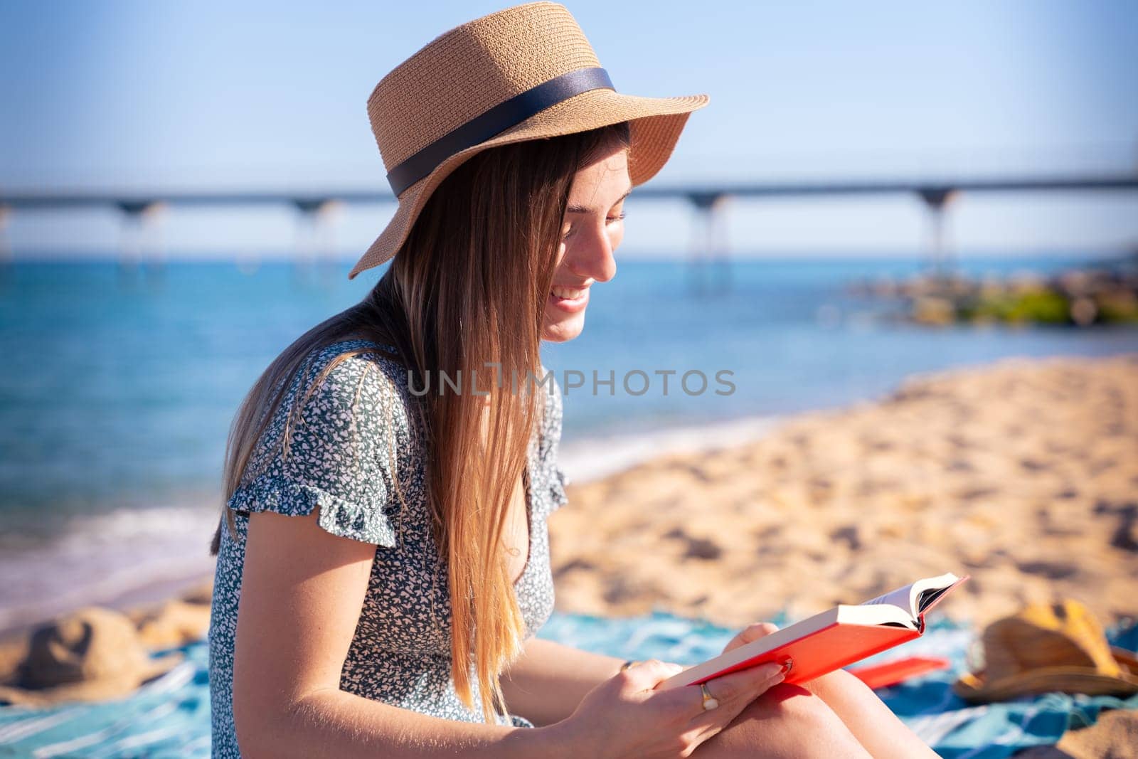 Woman sitting on the beach wearing hat reading a book,with the sea in the background
