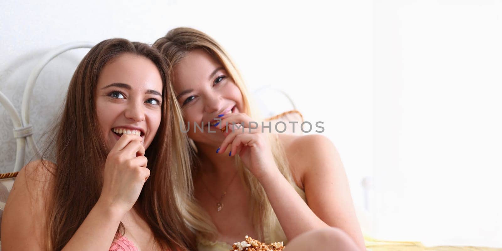 Portrait of beautiful young smiling girls friends watching comedy movie, eating popcorn and laughing on bed in pajamas. Friendship, lover, lgbt concept