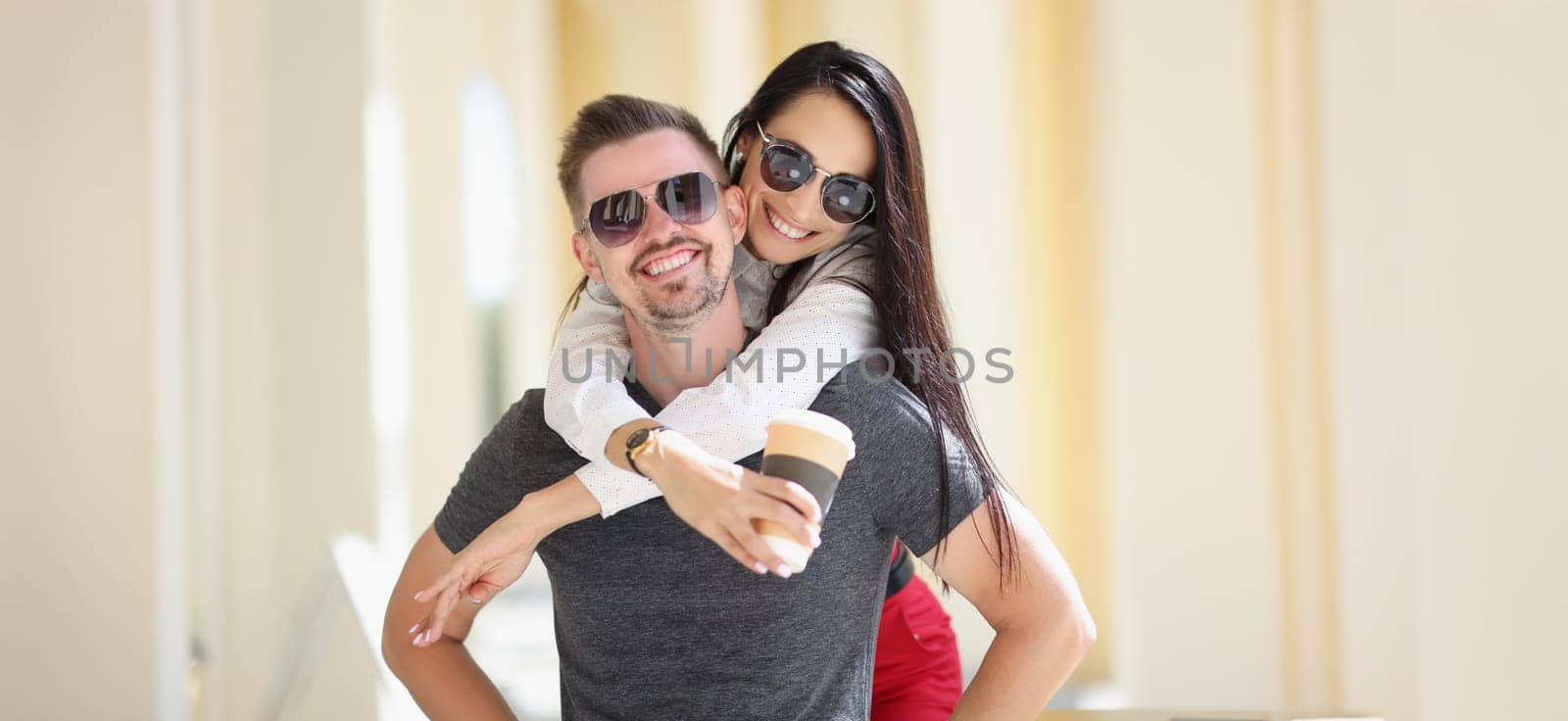 Portrait of happy couple posing, woman jumped on man back hold cup of coffee. Cheerful married people on vacation. Love, relationship, happiness concept