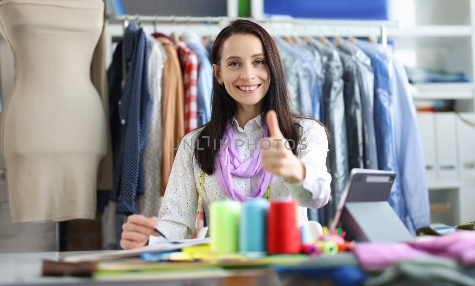 Young beautiful tailor woman showing thumbs up. Quality tailoring and clothing repair service concept