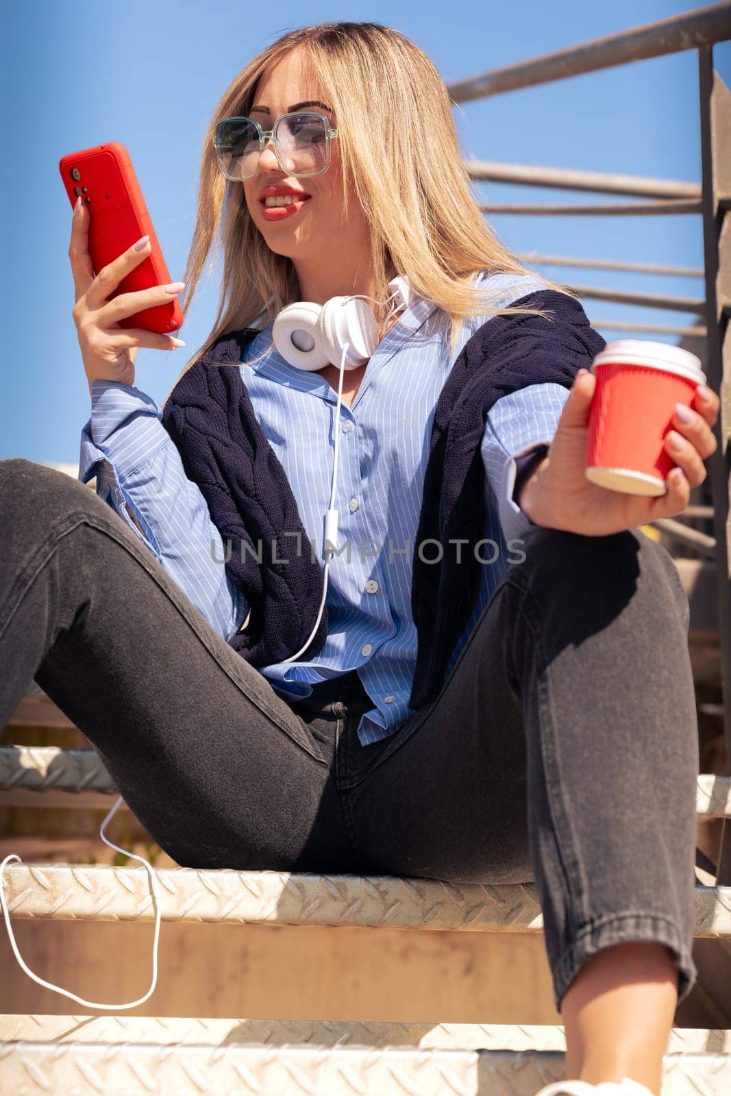 Portrait of a smiling young woman wearing headphones and sunglasses, using a social media app outdoors