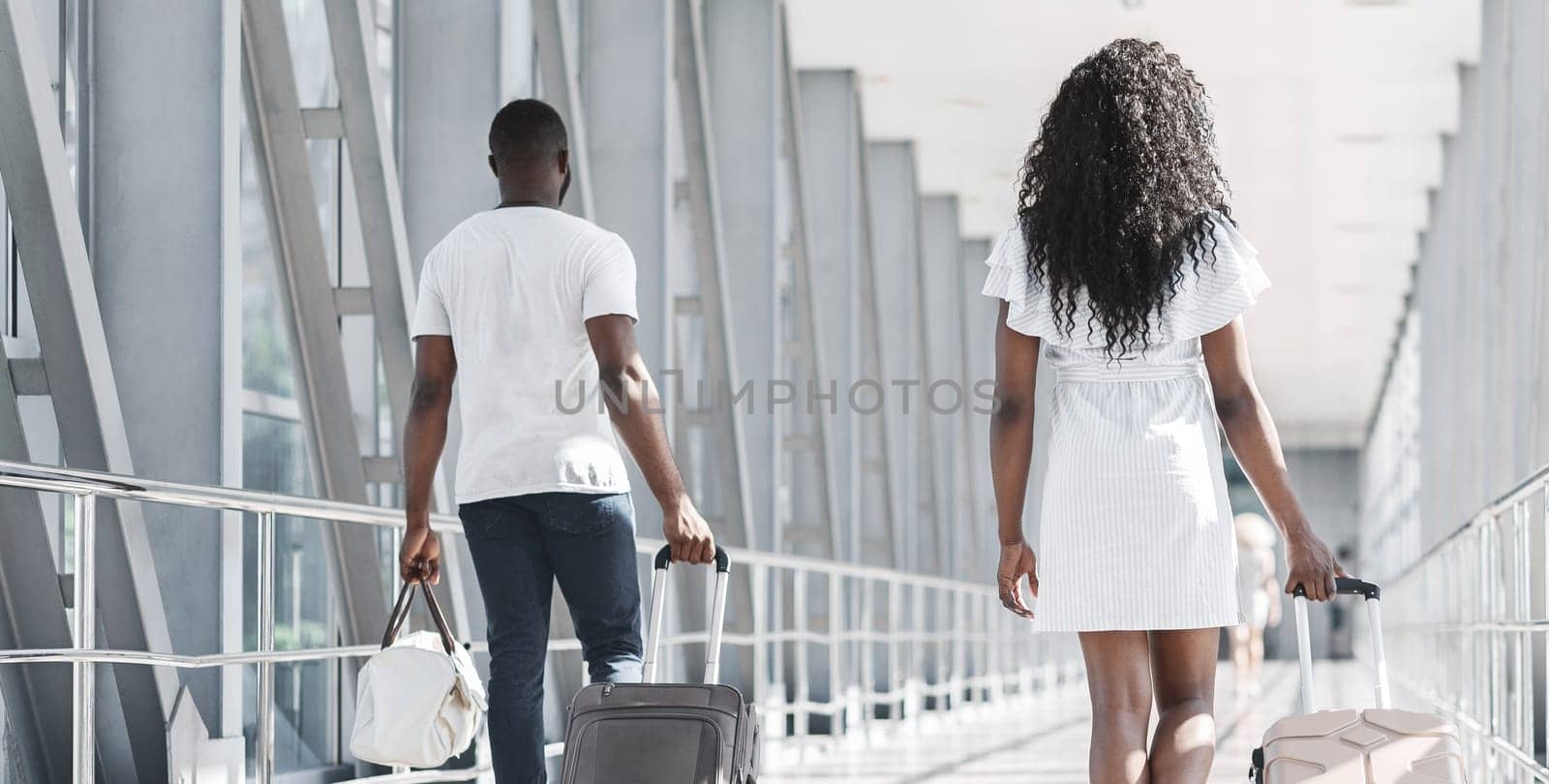 A couple walks through an airport concourse, each pulling a piece of luggage. The man is wearing a white shirt and jeans while the woman is wearing a white dress, back view