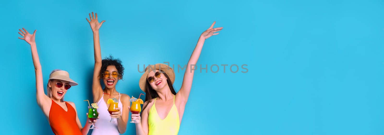 Three women wearing swimwear and sunglasses stand together in front of a blue background. They have their arms raised in the air, and each woman is holding a cocktail drink, copy space