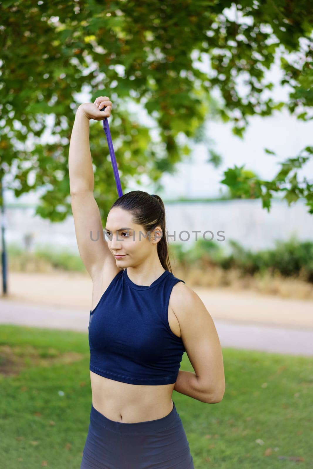 In a serene green park, an athletic woman in activewear is engaged in outdoor strength training using a resistance band. Fitness and wellbeing are essential in her workout routine