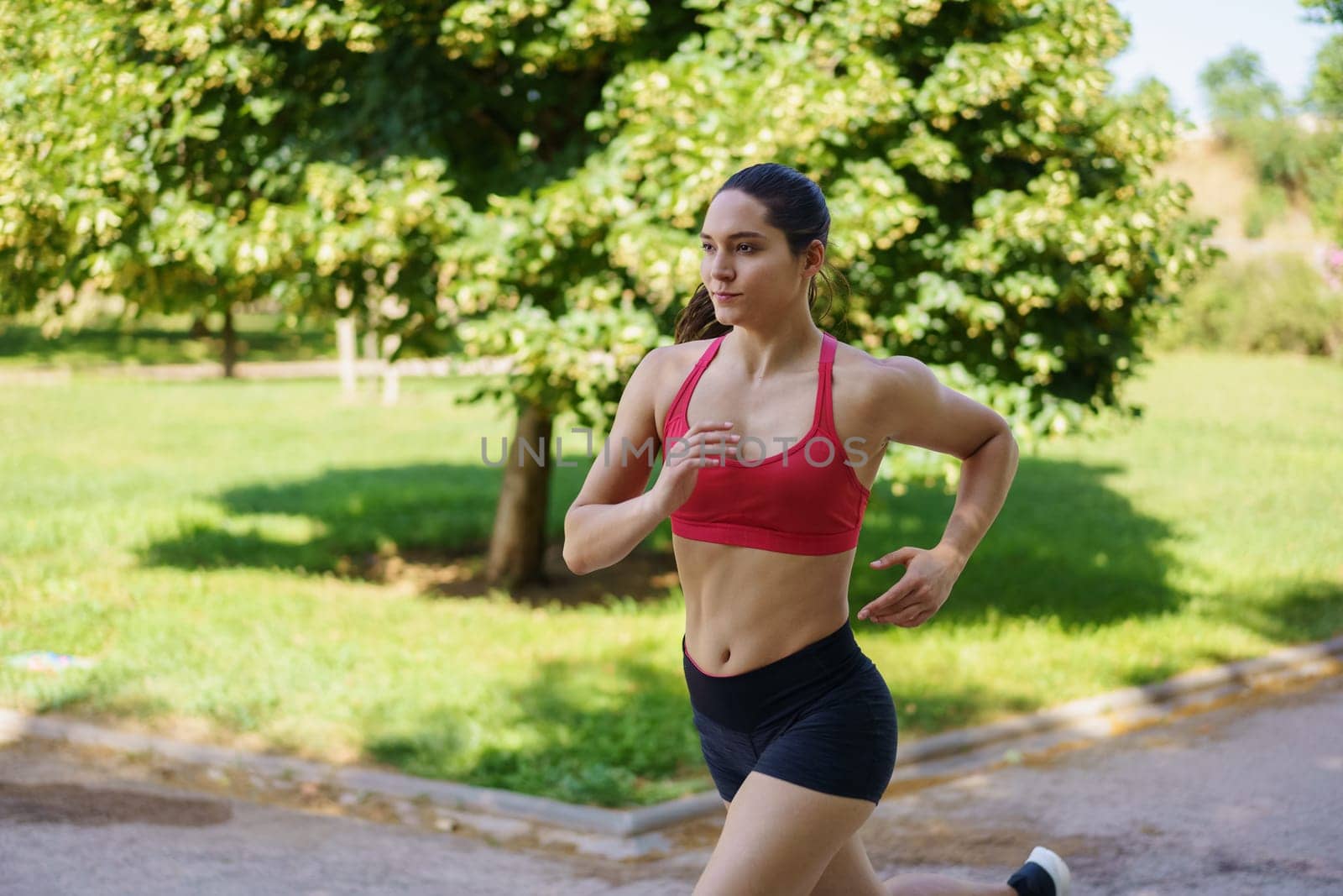 An active young woman is jogging in a park, embodying fitness and a healthy lifestyle. Dressed in a red sports bra and black shorts, she looks focused and determined in her workout