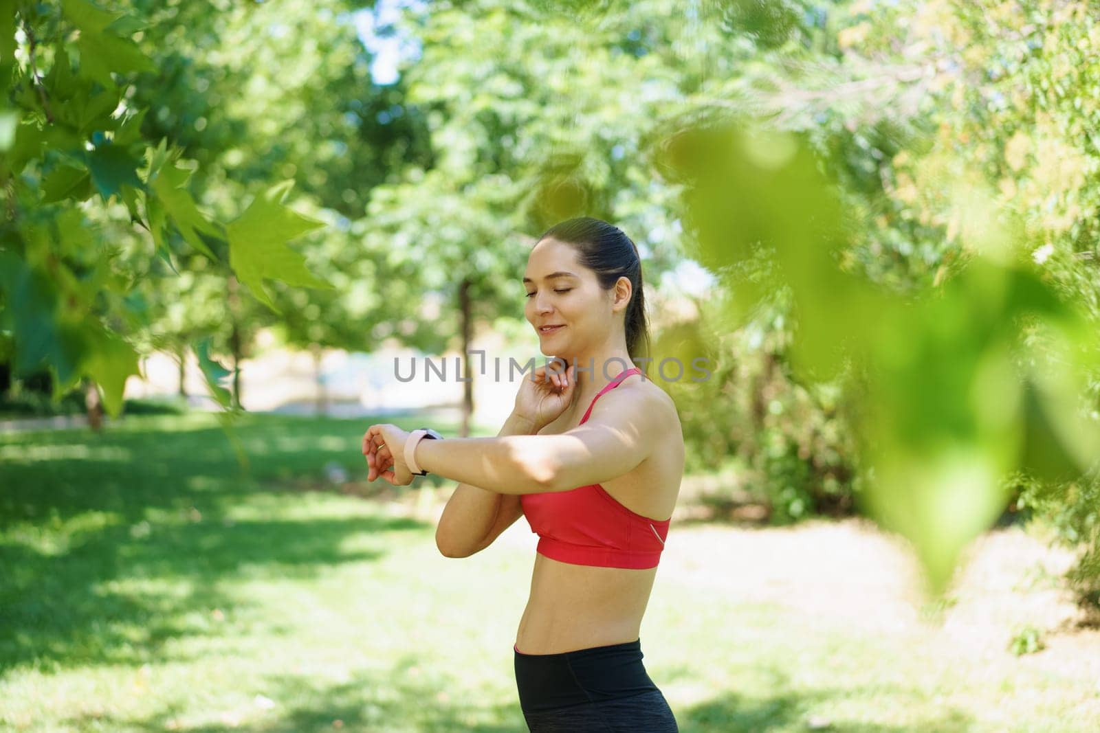 In a scenic green park, a young woman in sportswear uses a smartwatch to track heart rate and fitness progress while exercising, embodying dedication to an active lifestyle in nature