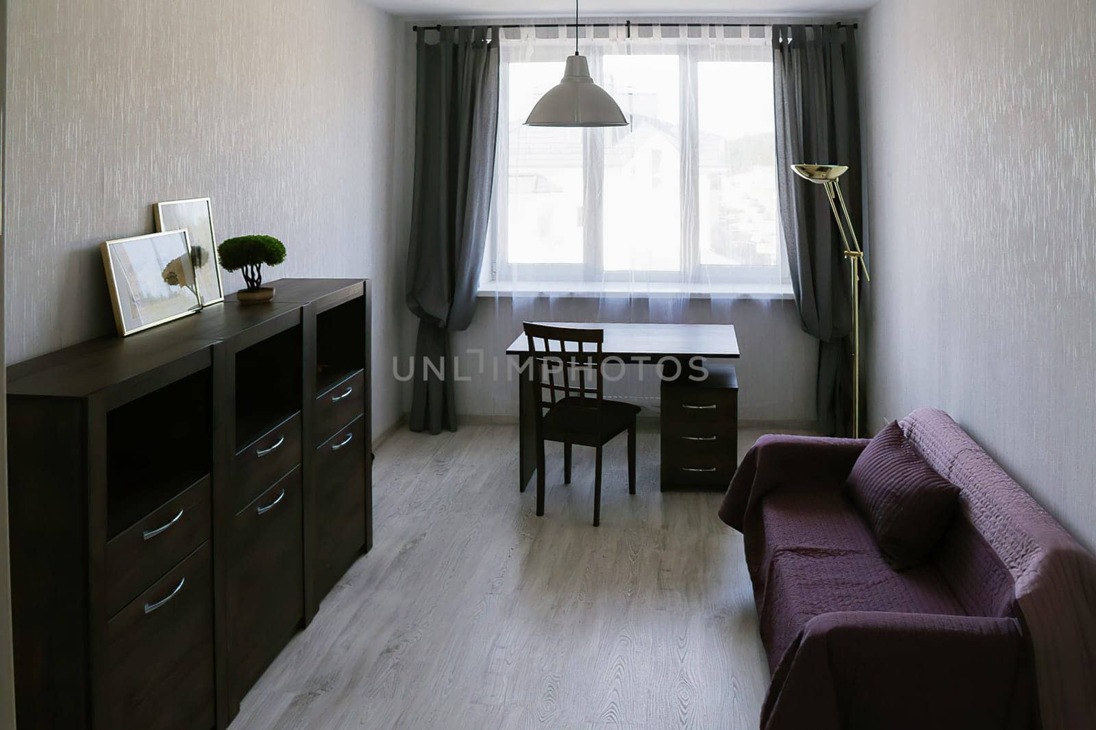 An office in an apartment consisting of a red sofa, a desk, a chair, a floor lamp, a chandelier, a chest of drawers, a small decorative tree in a pot, paintings, a light window.
