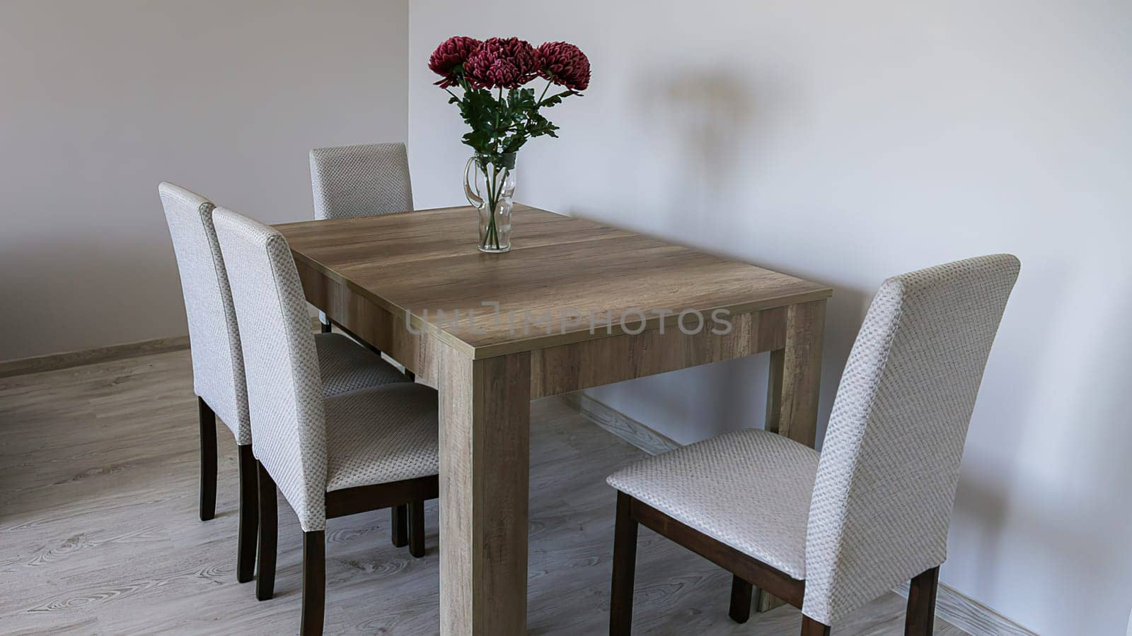 A brown wooden table, four fabric-covered chairs, a glass vase with three red flowers, light gray plain walls and laminate flooring.