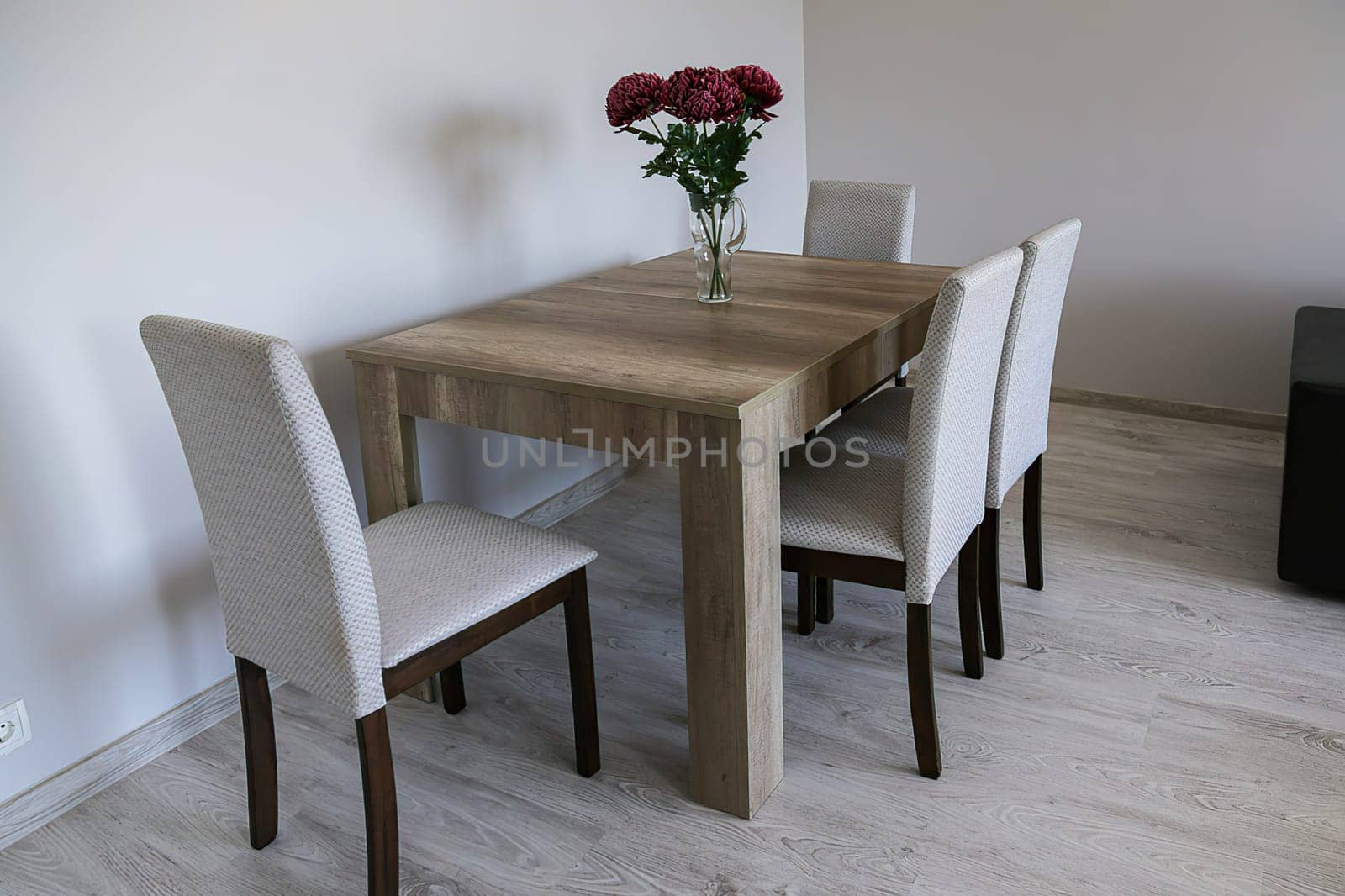 A brown wooden table, four light chairs, a glass vase with three red flowers, light gray plain walls and laminate flooring. Bright cozy room.