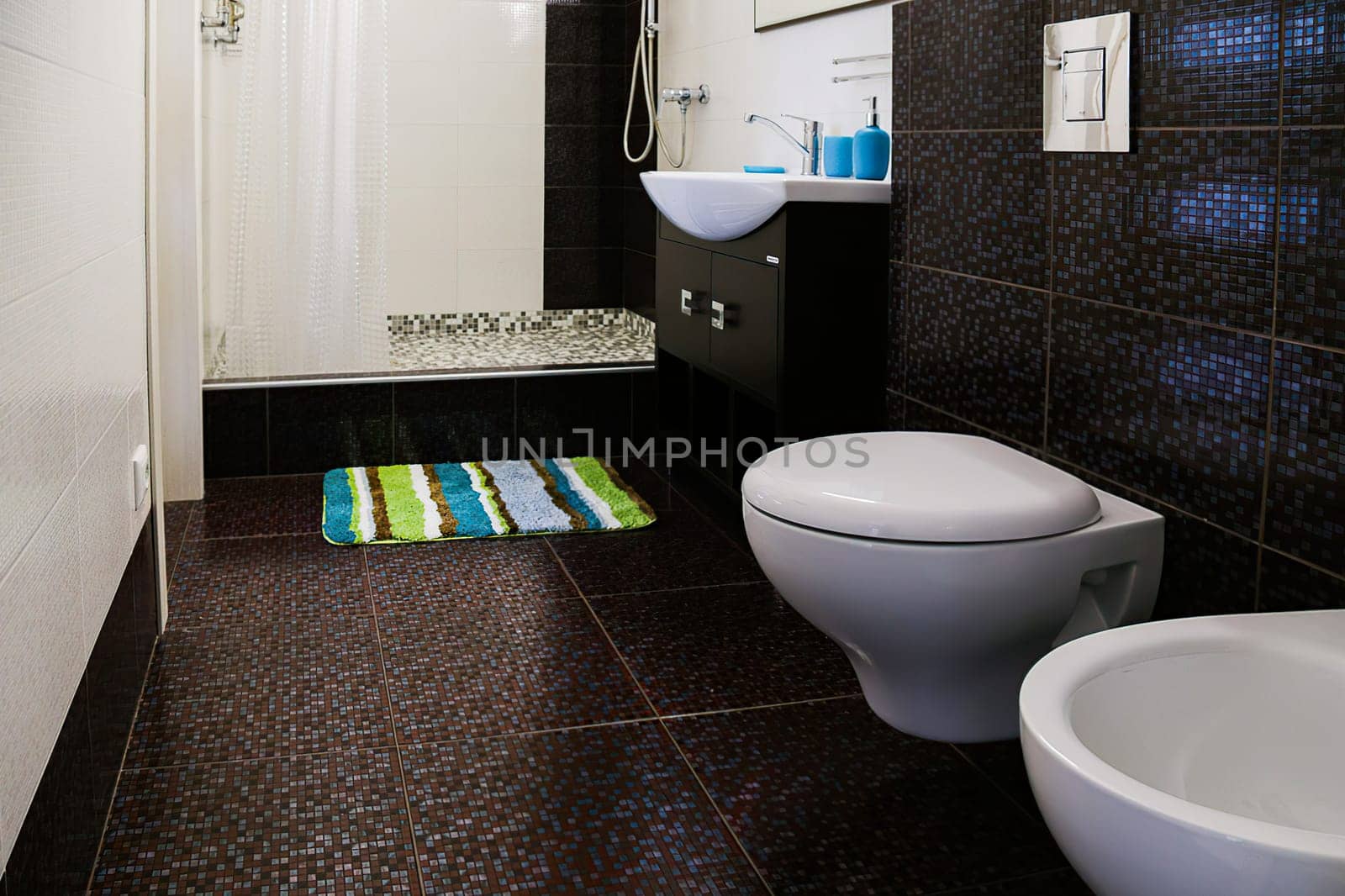 Interior of toilet with shower cubicle,floor and wall with brown tiles,sink,mirror,light fixture,floor mat,shower cubicle,curtains.