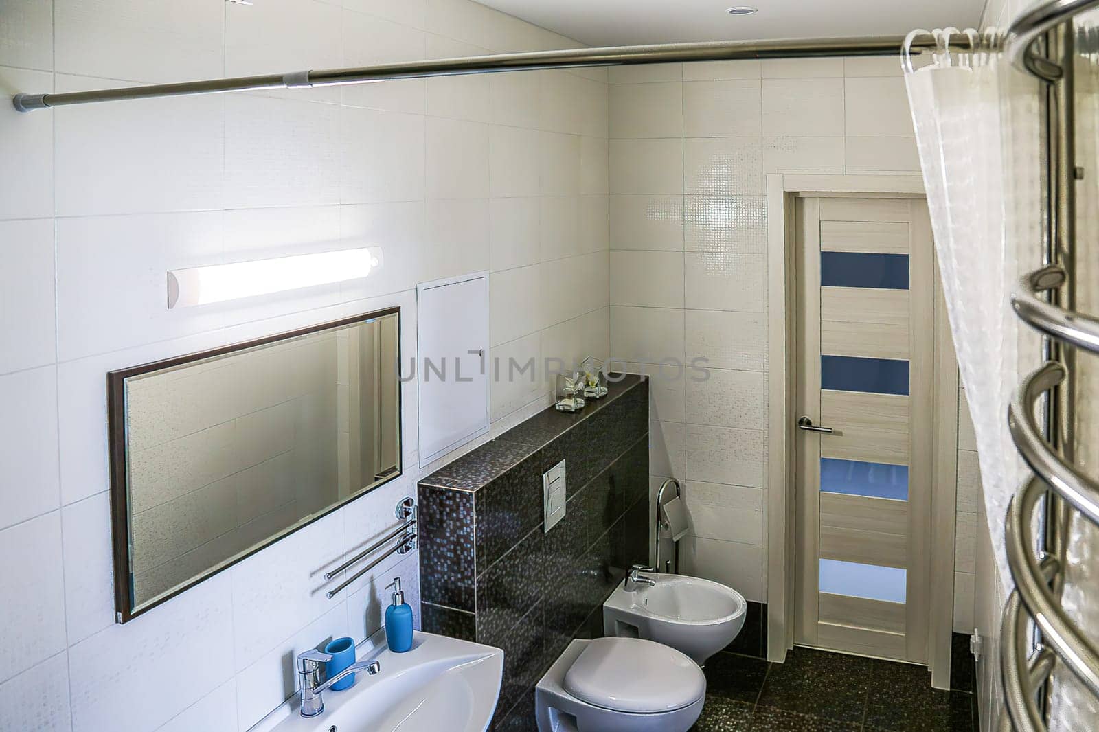 White toilet and bidet, dark brown tiled floor, light tiled walls, mirror, light fixture above mirror, sink with soap accessories, doors, red towel, towel dryer and rolled up shower curtain, toilet and bidet in bathroom,top view.
