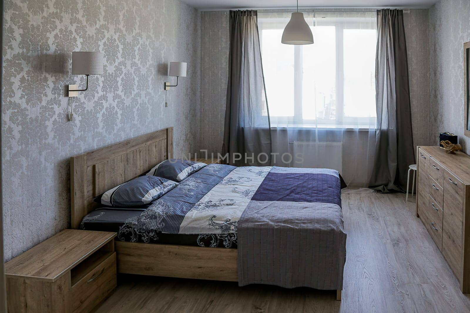 Interior of a small bedroom. Bedroom in a modern apartment with a double bed with a blue bedspread, a bedside table, a chest of drawers, two wall lamps, a chandelier, a window with open curtains.