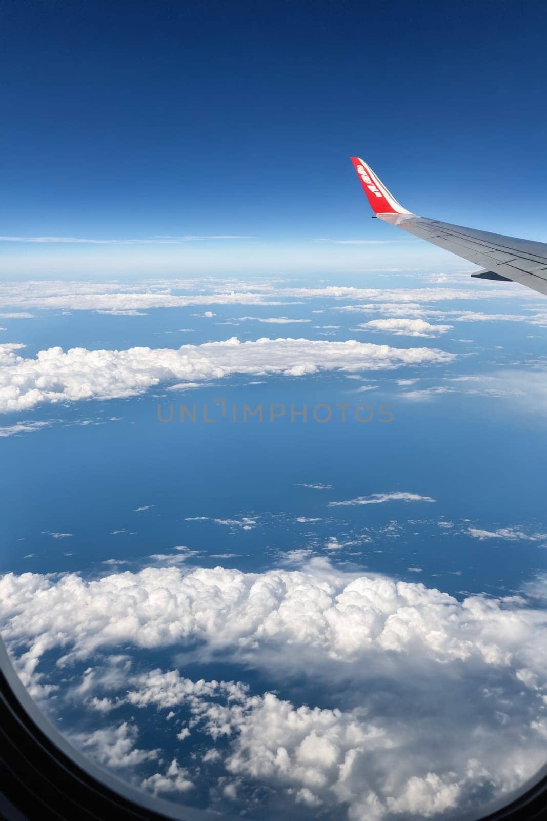 An airplane wing cuts through the clear blue sky, leaving behind a trail of white clouds. The view from the plane window showcases a breathtaking panorama of fluffy clouds scattered across the vast horizon. The bright blue sky and the pristine white clouds create a stunning contrast, making for a beautiful aerial landscape.