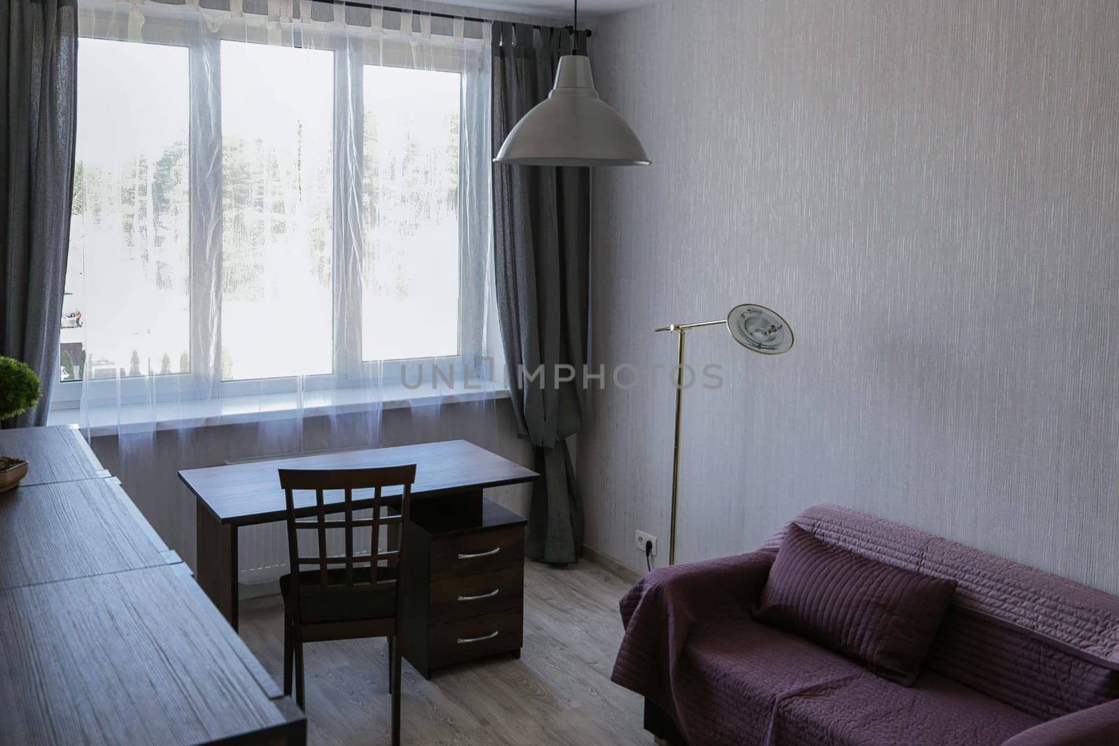 A small cozy office in a modern apartment with a sofa covered with a red blanket, a desk, a chair, a chest of drawers, a window, a floor lamp, and a chandelier.