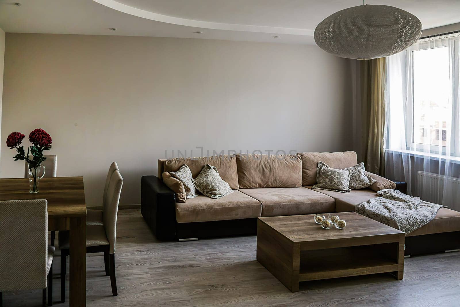 A cozy room with a large sofa with pillows and blankets, two tables, 4 chairs, a vase with red flowers, a window, a white round chandelier. Gray floor, walls, ceiling.