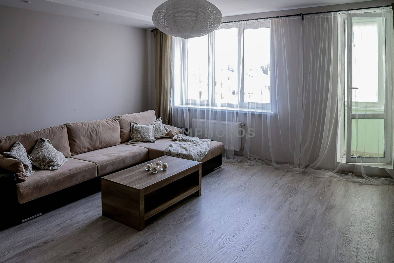 Large sofa with bedspreads and pillows, table, window, exit to the balcony, gray laminate flooring, white round chandelier.