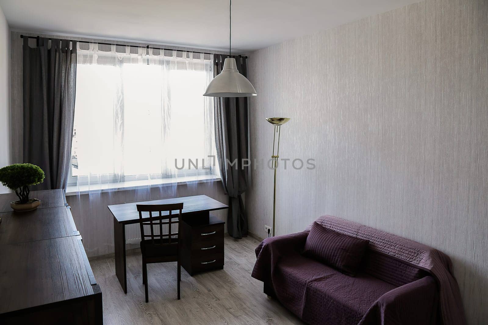 A small cozy office in a modern apartment with a sofa covered with a red blanket, a desk, a chair, a chest of drawers, a window, a floor lamp, and a chandelier.