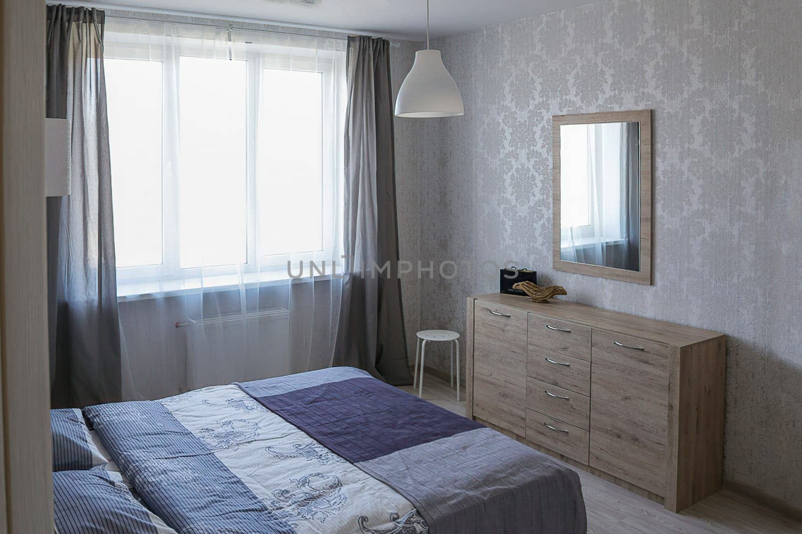 Interior of a beautiful bedroom. A bedroom in a new apartment with a double bed with a blue bedspread, a bedside table, a chest of drawers, two sconces, a chandelier, a mirror on the wall, a window with open curtains.