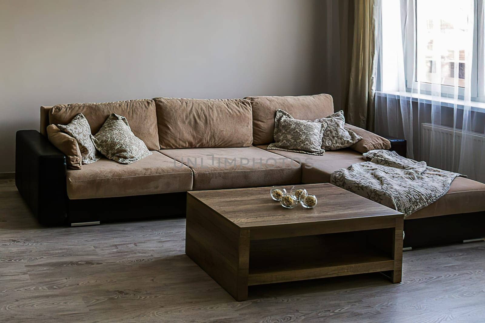 Fragment of a room with a sofa and table, a window with tulle. Living room with large sofa, pillows, coffee table, laminate flooring.