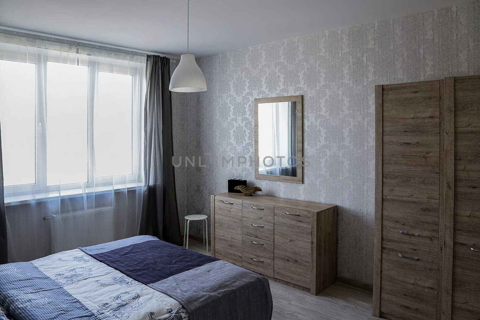 Double bed covered with a blue blanket, chest of drawers, wardrobe, mirror, window, white chandelier.