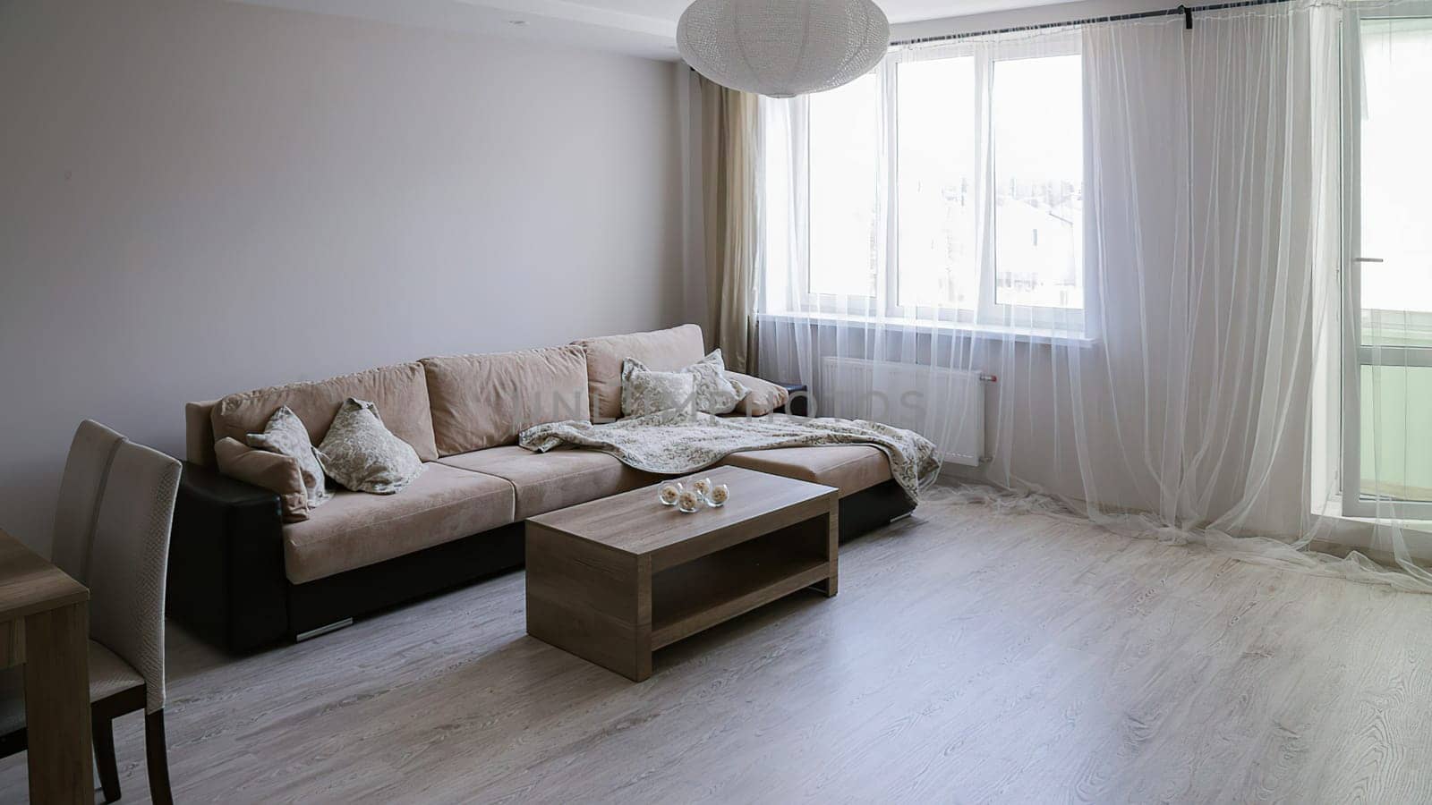 The interior of a large bright room with a corner sofa with bedspreads, two tables, two chairs, a window and doors to the balcony.