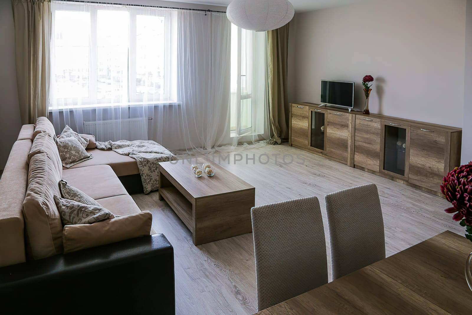 The interior of a large bright room with a corner sofa with bedspreads, two tables, two chairs, a chest of drawers with TV, a vase with a red flower.