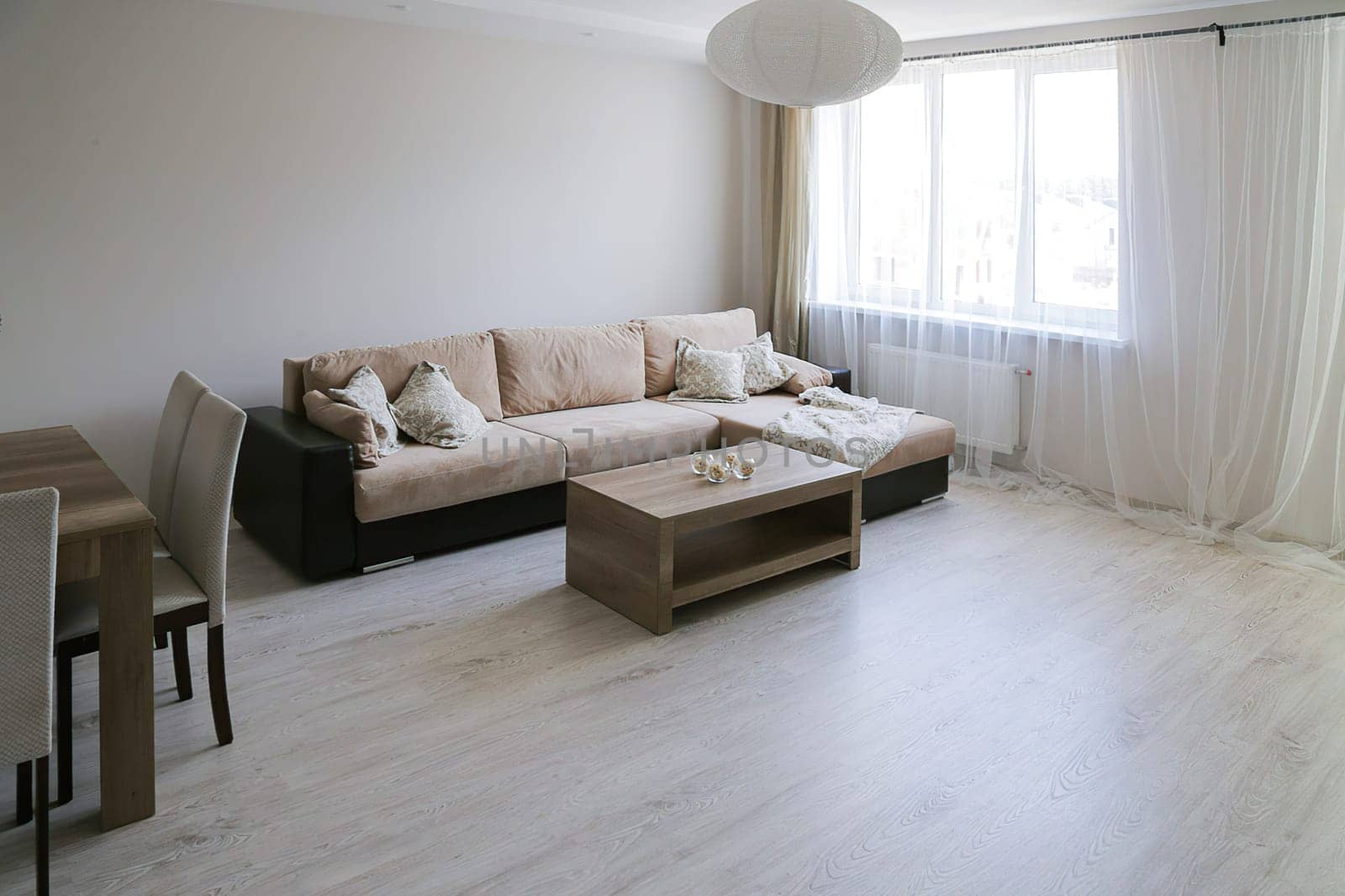 Bright, luxurious additional living room. Large corner sofa, coffee table, table with chairs, gray laminate floor, tulle window, white oval chandelier.