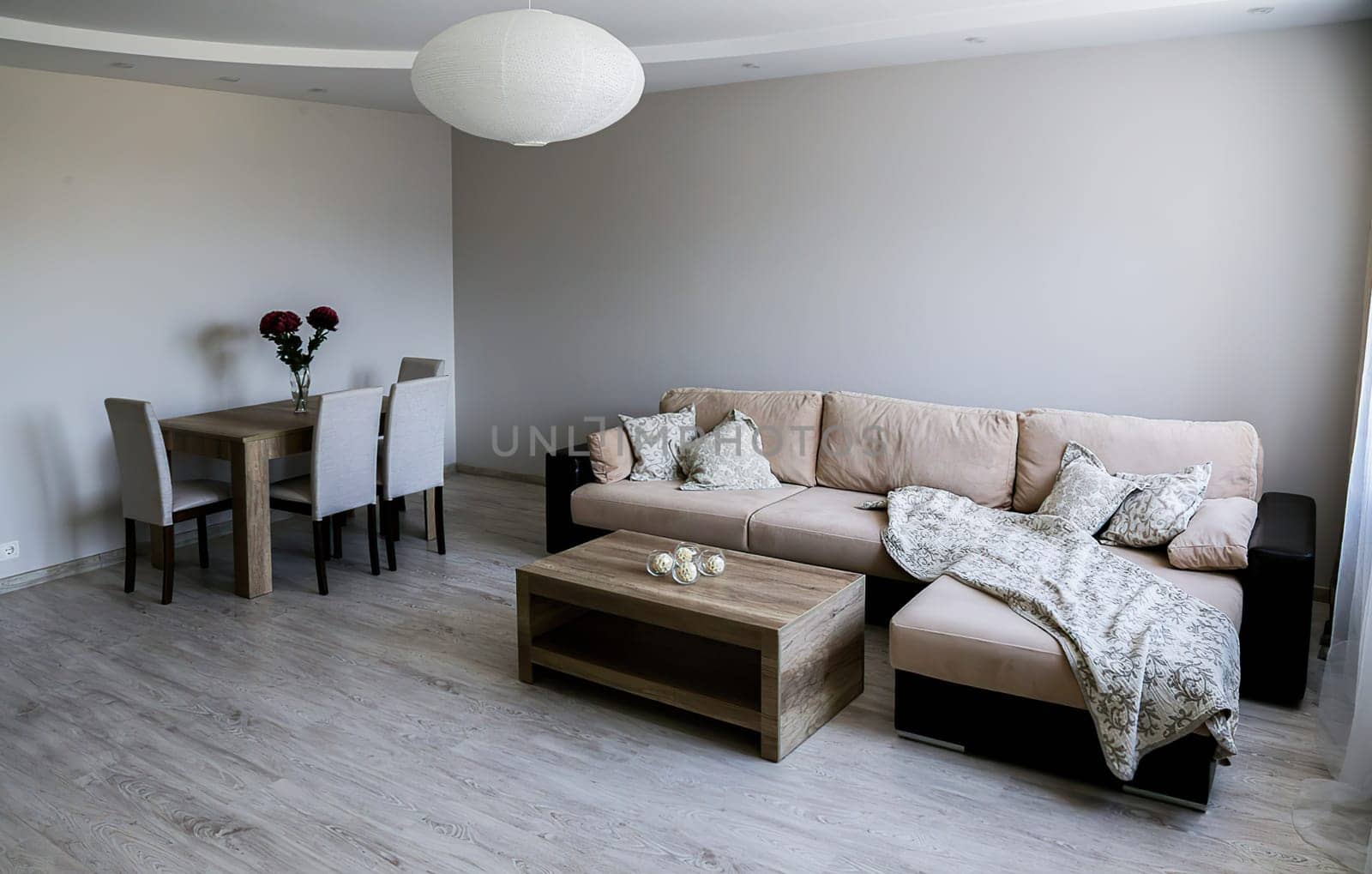 A cozy room with a large sofa with pillows and blankets, two tables, 4 chairs, a vase with red flowers, a white round chandelier. Gray floor, walls, ceiling.