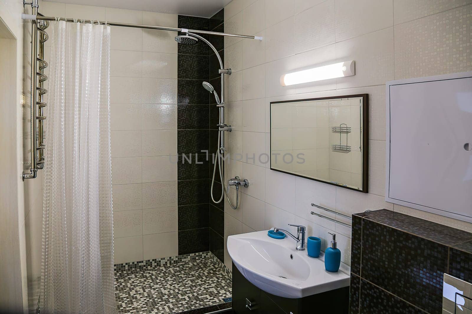 Interior of a modern minimalist bathroom. Sink is located under the mirror, lamp above the mirror, blue soaps on the sink, and shower cubicle with curtains.
