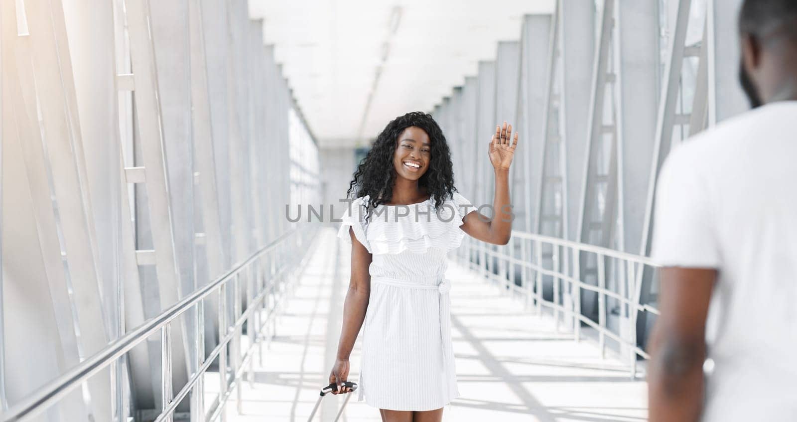 African American woman in a white dress walks down a terminal corridor at an airport, waving goodbye to someone out of frame. She is smiling and appears to be happy to be leaving.