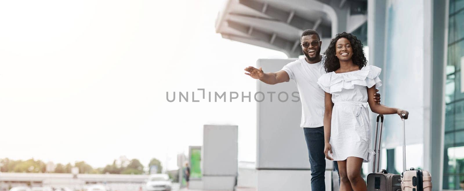 A smiling black couple arriving at an airport, pulling their luggage behind them, hailing taxi. The background is a modern airport terminal with large glass windows, copy space