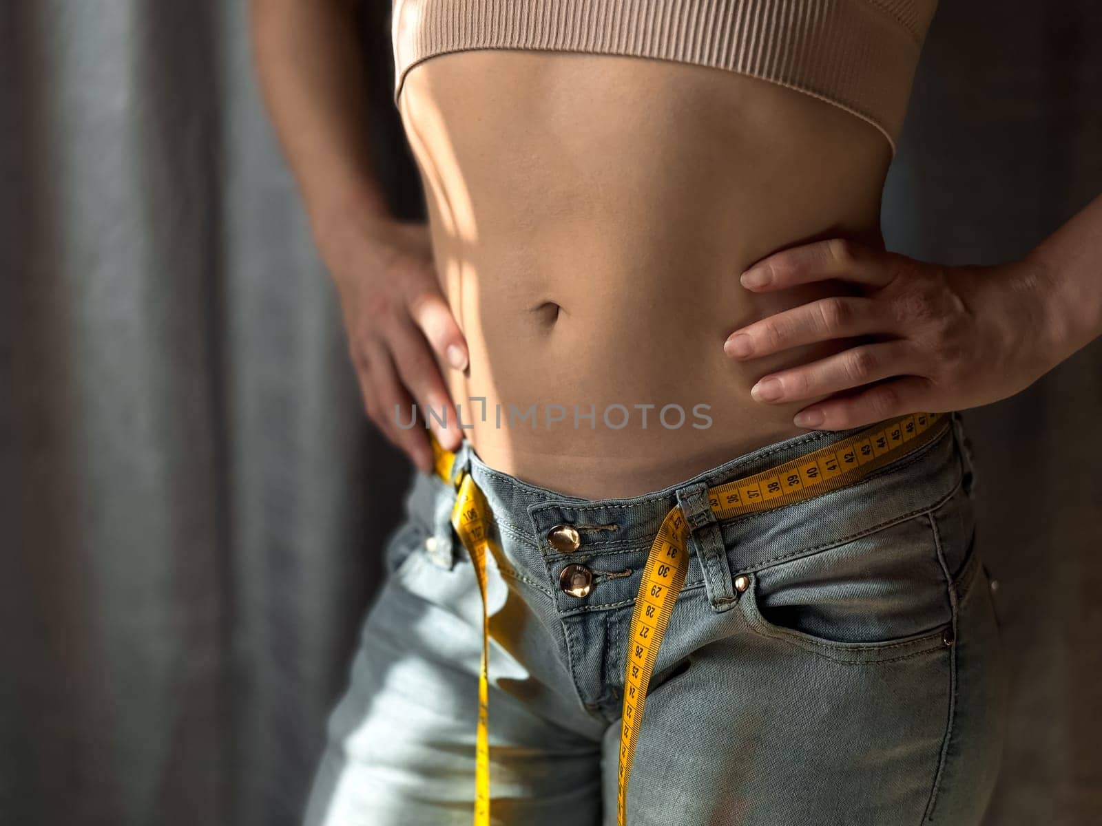 Close up of the naked torso of faceless woman in jeans with yellow measuring tape instead of belt on neutral background. Weight loss, fitness or body measurement concept. Lifestyle photo. High quality