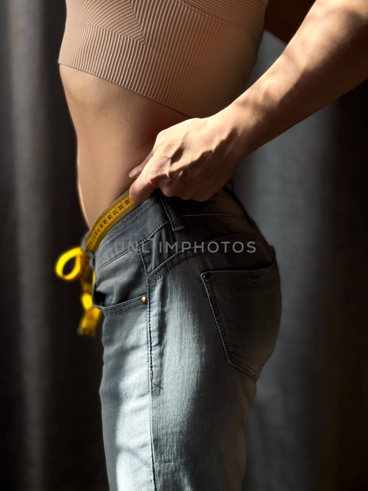 Close up of the naked torso of faceless woman in jeans with yellow measuring tape instead of belt on neutral background. Weight loss, fitness or body measurement concept. Lifestyle photo. High quality