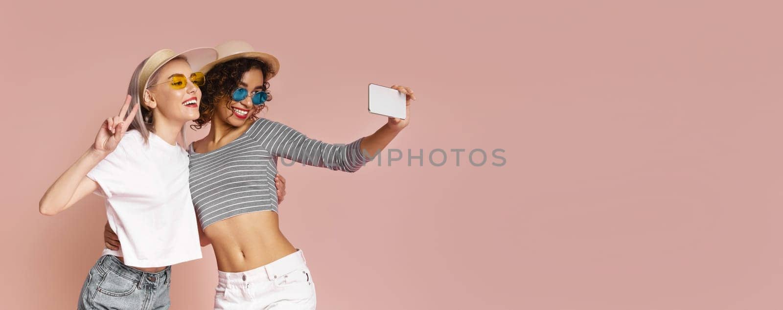 Two women, one wearing a white shirt and the other a striped shirt, are taking a selfie photo against a light pink background. They are both smiling and wearing sunglasses and hats.