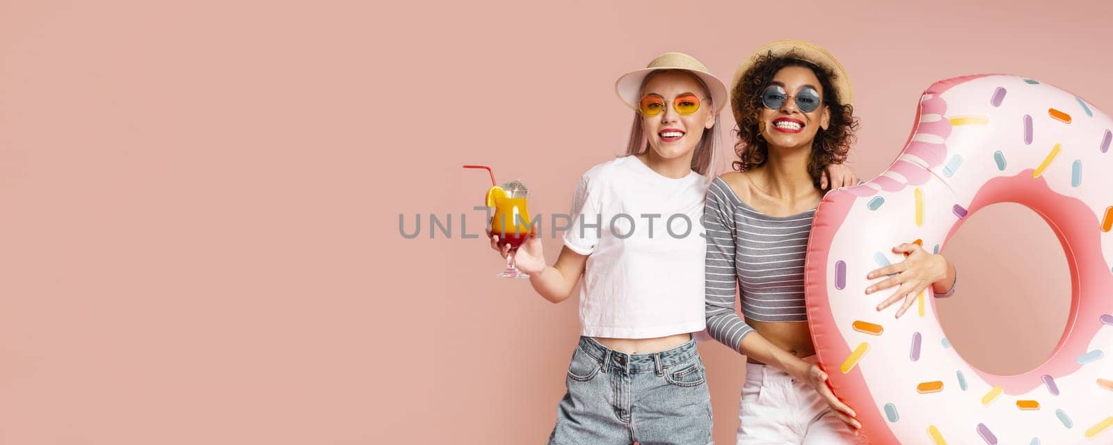 This image shows two happy friends posing with a donut pool float on a pink background. Both are smiling and wearing sunglasses. This image is perfect for representing summertime fun and friendship.