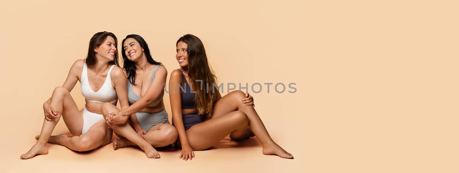 Three glad diverse millennial women in undergarments sit on floor, enjoy body positivity, beauty care, have fun, isolated on light studio background, full length. Friendship, confidence, copy space