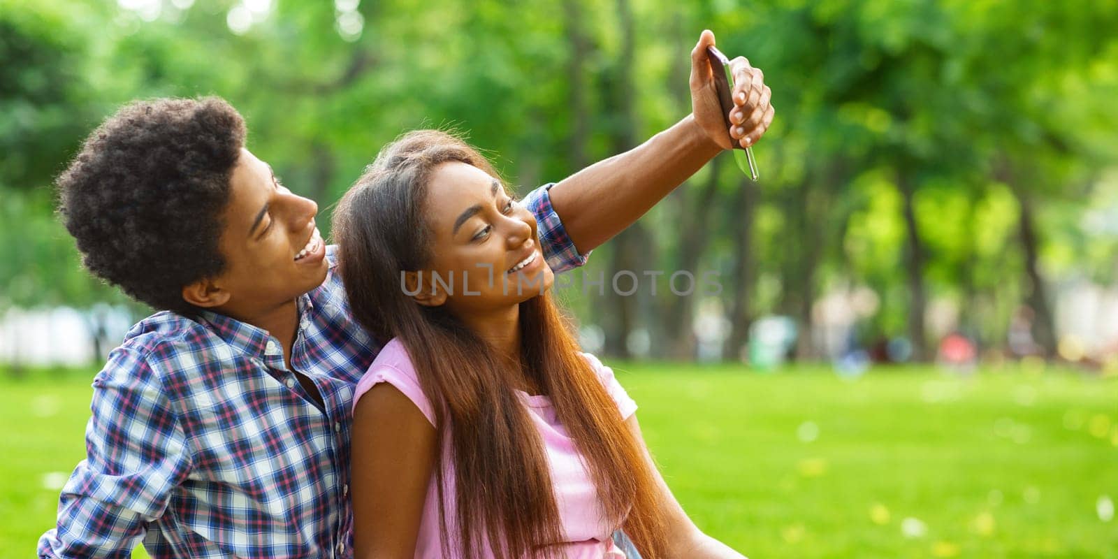 A teen black couple sitting on a grassy field in a park on a sunny day, taking selfie on smartphone. The couple is enjoying the beautiful weather and each others company, copy space