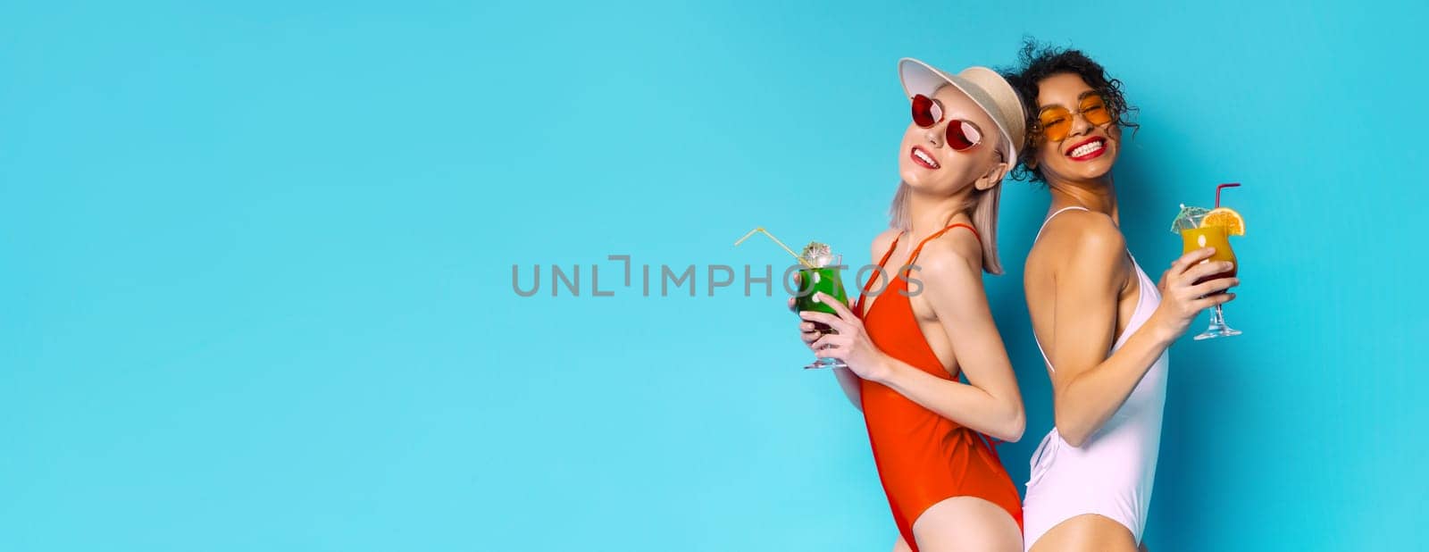 This image shows two women in swimsuits, one wearing a white swimsuit and the other wearing a red swimsuit, smiling and holding cocktails against a bright blue background, copy space