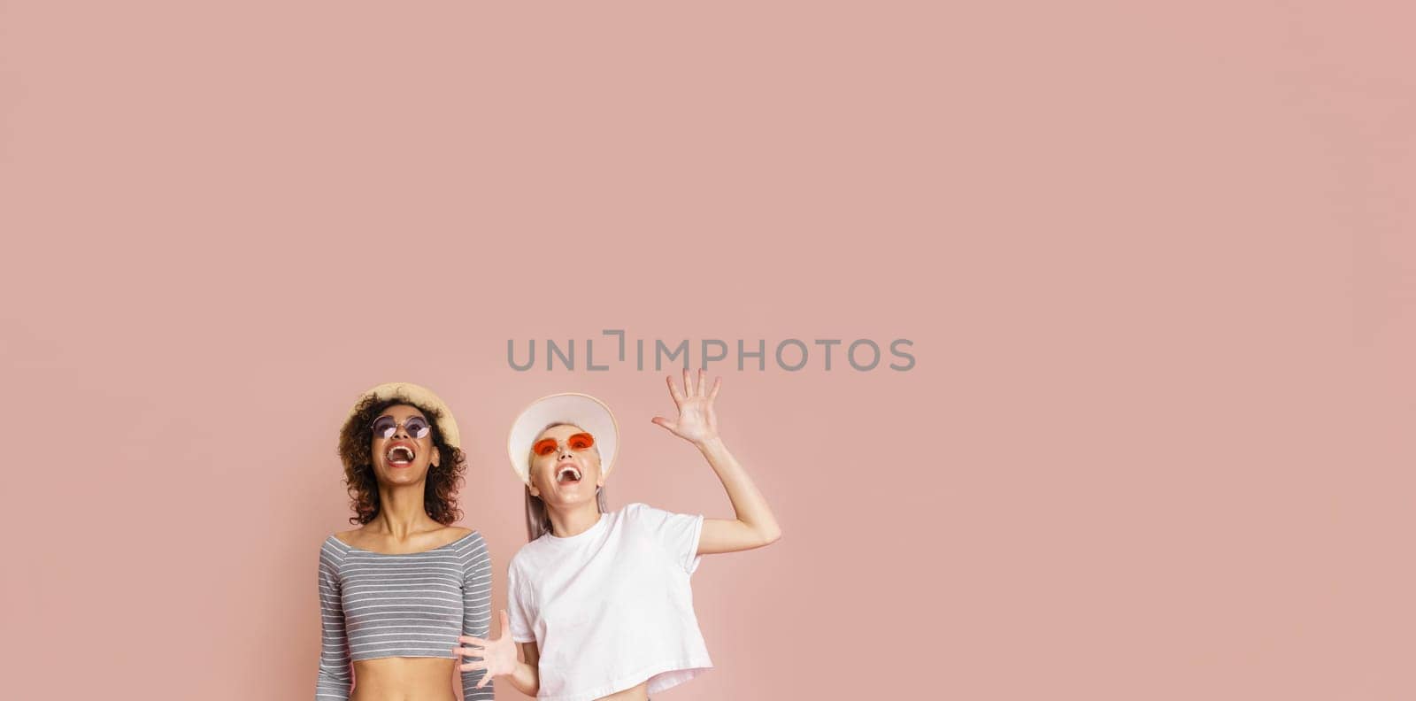 Two young women are laughing at the camera while wearing sunglasses and hats. The background is a solid pink color, copy space