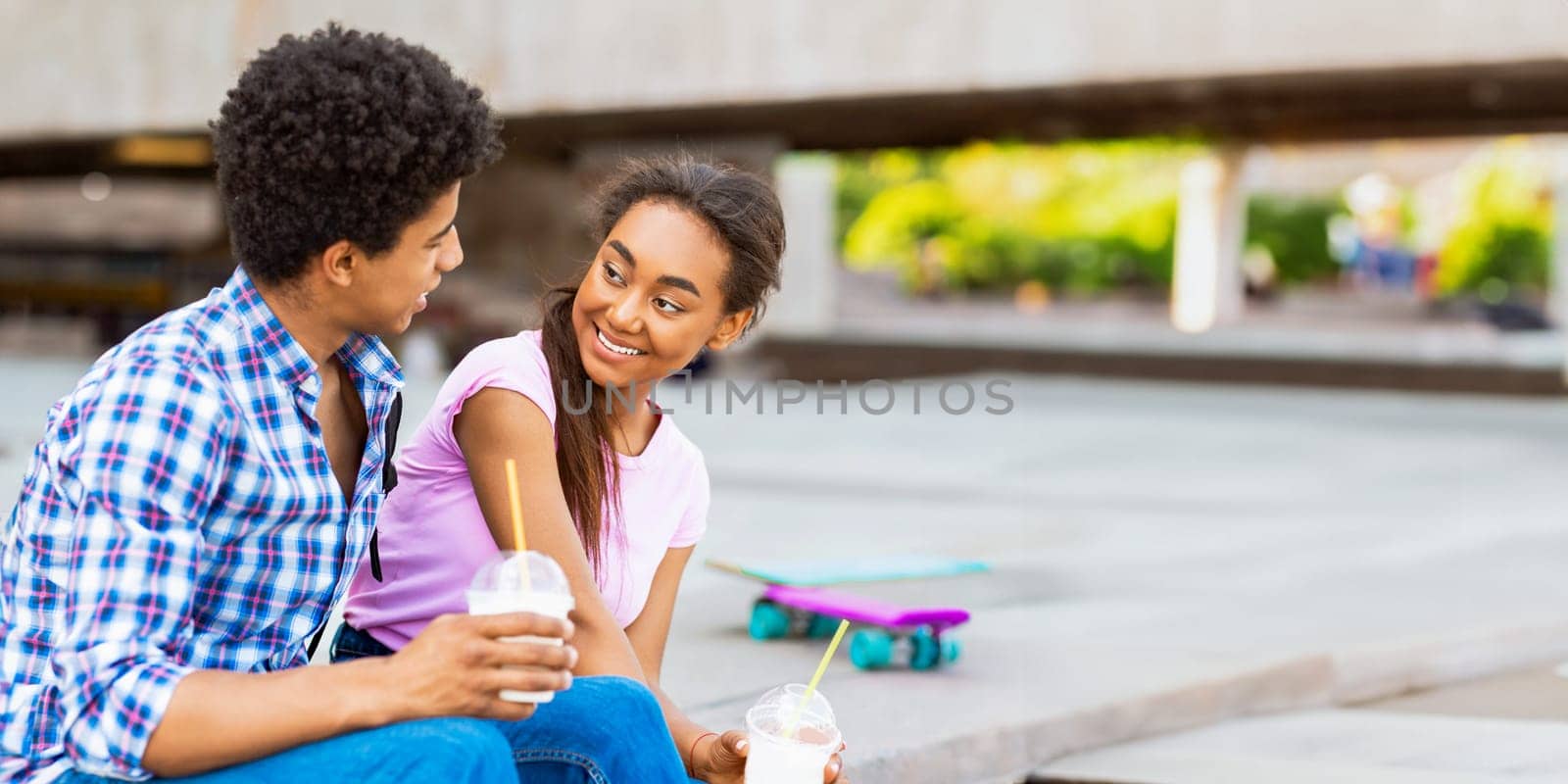 A teen African American couple sits together on a concrete ledge, both holding drinks with straws. They are engaged in conversation, smiling at each other, and enjoying a sunny day, copy space