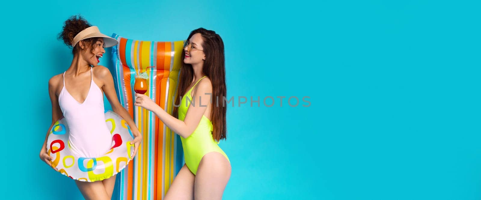 This image depicts two young women, one in a white swimsuit and the other in a green swimsuit, standing together in front of a bright blue background, copy space